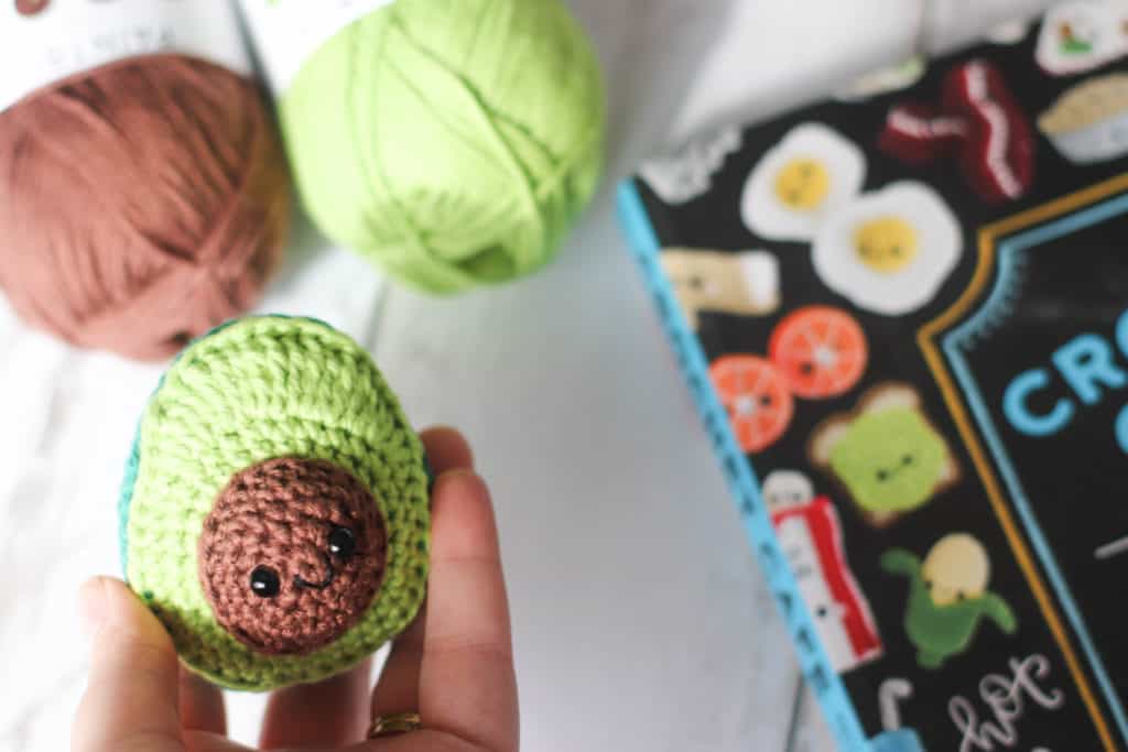 A crocheted avocado amigurumi with the crochet cafe book and brown and green yarn in the background.