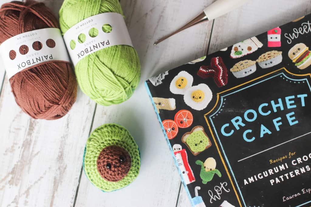 A crocheted avocado amigurumi with the crochet cafe book and brown and green yarn in the background.