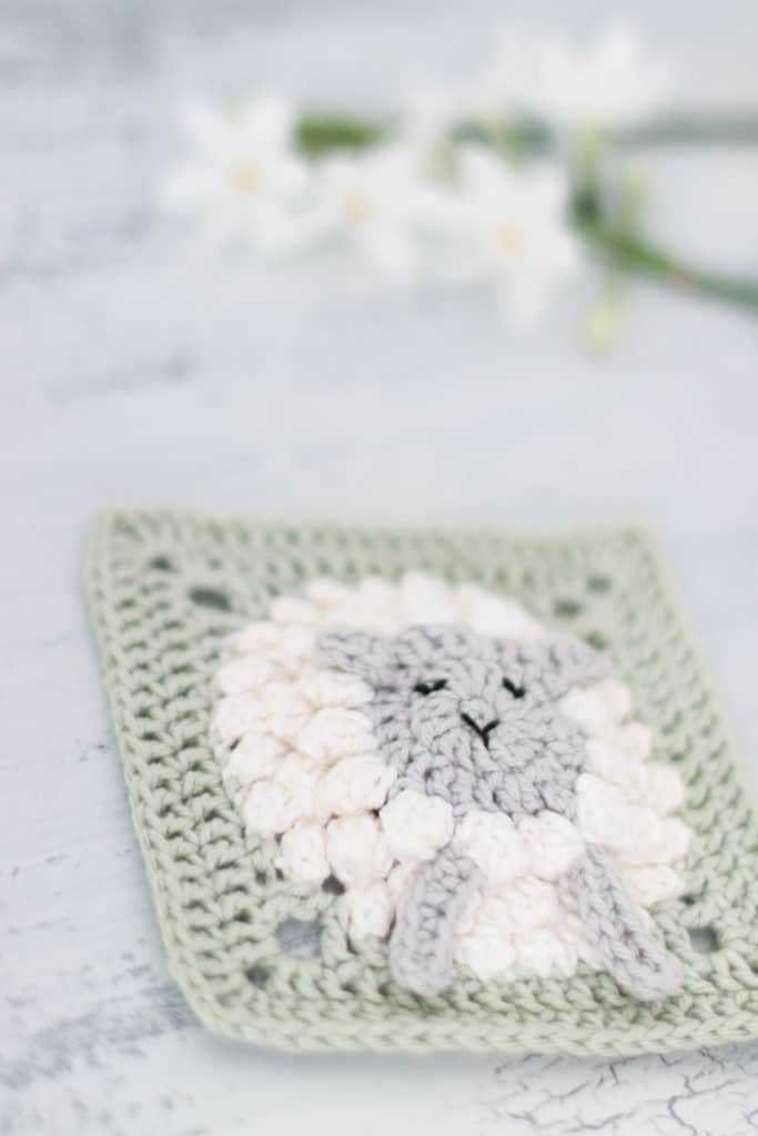 Fluffy white lamb, green crocheted square with flowers in the background. 