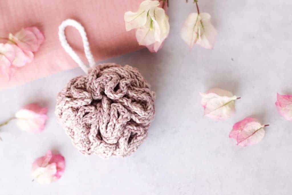 pink crocheted shower pouf on a granite background surrounded by soft pink fabric and flowers, spa scene