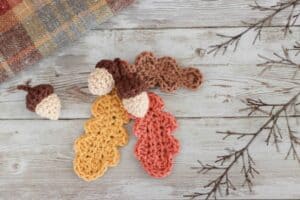 crochet acorns and crochet oak leaves on a wooden background surrounded by twigs and tartan