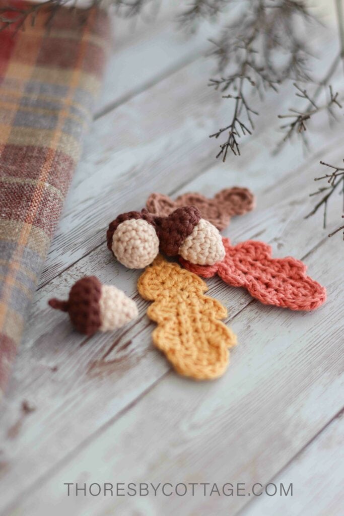 crochet acorns and crochet oak leaves lying on a wood textured background, surrounded by twigs and tartan