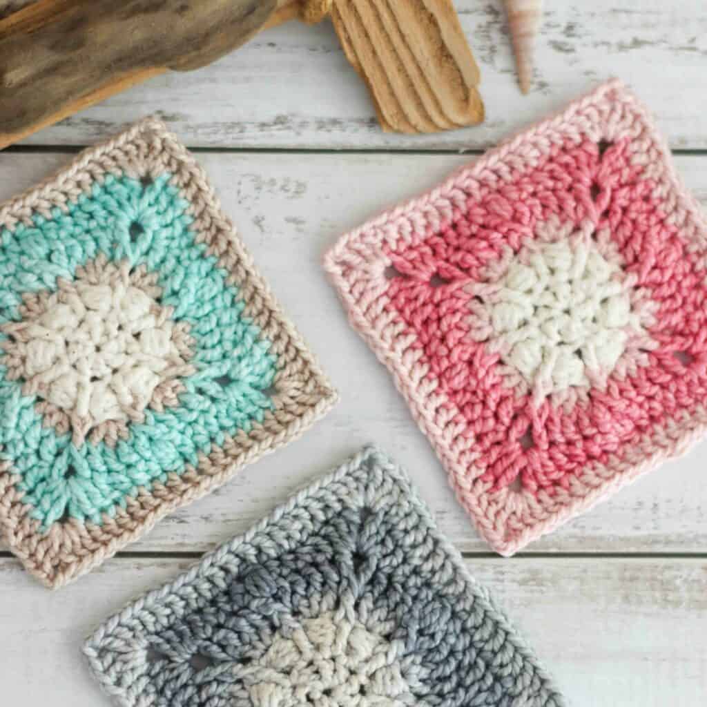 Sand dollar crochet square pattern, in blues, greys and shades of pink. The crochet squares are on a wooden textured background.