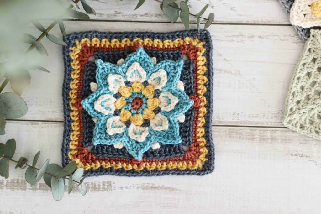 Lotus flower granny square in rich jewel tones, surrounded by penny gum branches on a wooden textured background.