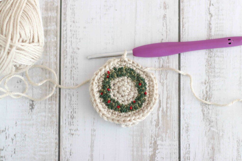 Crochet wreath Christmas ornament, a green wreath embroidered onto a circle of crochet. This ornament is laid on a wooden background surrounded by yarn and a crochet hook.