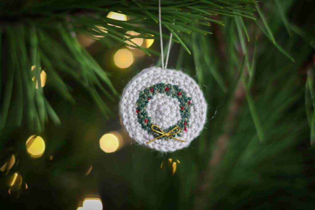 Embroidered crochet Christmas wreath ornament hanging in a Christmas tree, surrounded by twinkling lights.