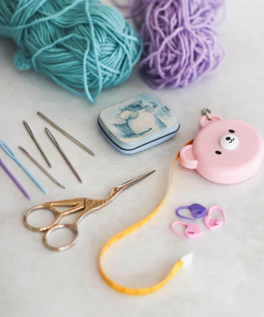 essential crochet supplies: yarn, measuring tape, tin of pins, sewing needles and stitch markers