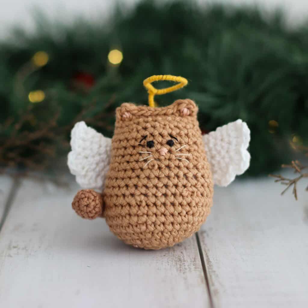 crochet angel cat pattern with white wings and a halo, surrounded by green foliage.